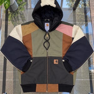 May include: A color-blocked Carhartt hooded jacket featuring panels of olive green, navy blue, tan, brown, and pink. The jacket has a full-length front zipper, a black quilted hood with a cream-colored top panel, and a Carhartt logo patch.