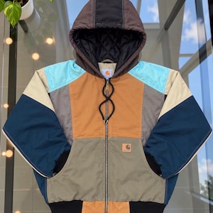May include: A color-blocked hooded jacket featuring a brown, navy blue, light blue, tan, and olive green panel design. The jacket has a full-length front zipper, a quilted black hood lining, black ribbed cuffs, and a Carhartt brand patch.