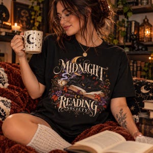 May include: A black t-shirt featuring a graphic print of a black cat resting on an open book with the text Midnight Reading Society, surrounded by floral elements and a crescent moon. The shirt is paired with a cream-colored mug with moon motifs.