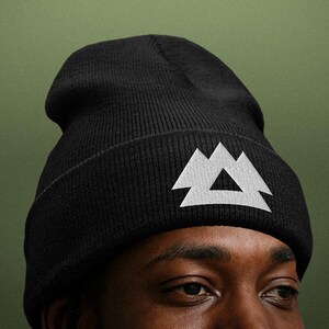 May include: Black beanie with a white geometric mountain design. The hat is made of a knitted material and is worn on a person's head. The background is a solid olive green.