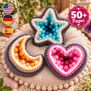 May include: Three crocheted plush items in the shapes of a star, a crescent moon, and a heart. The star is filled with blue beads, the moon with orange and yellow beads, and the heart with pink beads. Each item has a dark gray crocheted border.