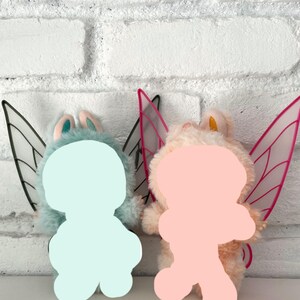 May include: Two plush fairy dolls. One doll is light blue with black and white wings, and the other is peach with pink and white wings. Both dolls have a light pink face and ears.