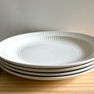 May include: A stack of four round, off-white ceramic dinner plates. Each plate features a smooth, flat centre surrounded by a wide, raised rim decorated with a series of fine, vertical, radiating grooves that extend to the outer edge.