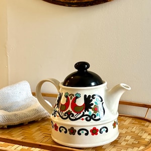 Puede incluir: Una tetera de cerámica blanca con tapa negra y un patrón gráfico colorido. El diseño incluye dos pájaros rojos con detalles en verde, rodeados de volutas negras y un borde inferior con motivos florales en rojo, verde y azul.