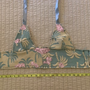 May include: A blue and beige fabric top with a palm tree print and pink sequins. The top has two straps and measures 17 inches across.