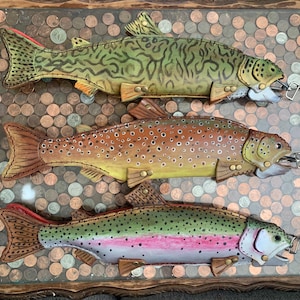 May include: Three handcrafted leather fish-shaped zipper pouches. The top pouch is olive green with dark brown mottled markings. The middle pouch is tan with brown and white spots. The bottom pouch is silver with a pink side stripe and black spots.