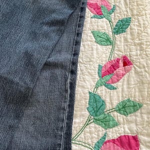 May include: Blue denim jeans with a classic five-pocket design, partially covering a white quilt. The quilt features a floral pattern with pink roses and green leaves, adding a touch of color.