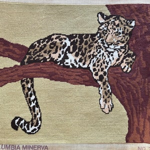 May include: A needlepoint tapestry featuring a leopard resting on a tree branch. The leopard has a spotted pattern in shades of brown, black, and white. The tree branch is brown, and the background is a light tan color. The text "COLUMBIA MINERVA" is at the bottom.