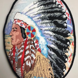 May include: A needlepoint pillow featuring a Native American man wearing a headdress with a white and blue feather design. The man is wearing a brown and red patterned shirt and a tan and brown fur coat. The background is a blue and white sky with mountains in the distance.