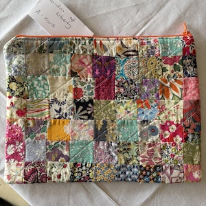 May include: A rectangular quilted pouch featuring a patchwork design of various floral and patterned fabric squares in shades of pink, blue, yellow, green, and black. The pouch has an orange zip closure and a white tag labelled London of Liberty A - Front.