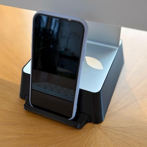 May include: A black smartphone with a light-coloured protective case resting in a black plastic charging dock. The dock features a silver-coloured top surface with a leaf-shaped cutout and is positioned directly in front of a silver computer monitor stand.