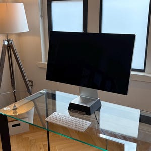 May include: A computer monitor with a black screen and a silver stand sits on a glass desk. A white wireless keyboard is placed on the desk surface in front of the monitor. The monitor base is black and rectangular with a silver metallic neck.