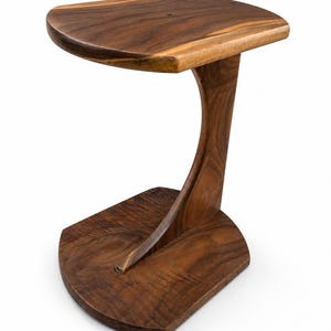 May include: A C-shaped side table crafted from dark brown wood with a visible grain pattern. The table features a flat, rounded rectangular top supported by a single curved vertical post that connects to a matching flat, rounded rectangular base.