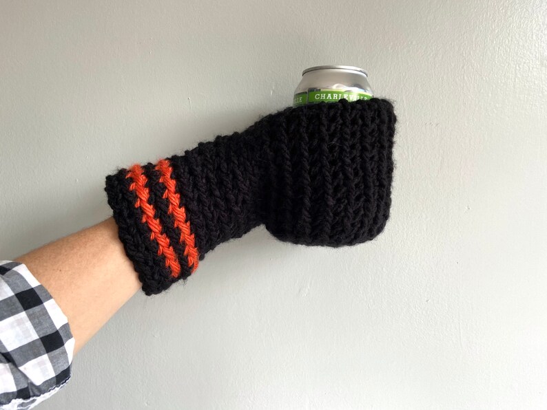 This beer mitten is black with orange gold stripes around the wrist. It is part crochet can holder part mitten. A hand is in it and a can of beer inside the holder. The bottom is covered so the can stays in place. It can be used on either hand.