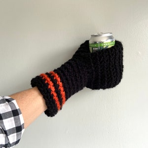 This beer mitten is black with orange gold stripes around the wrist. It is part crochet can holder part mitten. A hand is in it and a can of beer inside the holder. The bottom is covered so the can stays in place. It can be used on either hand.