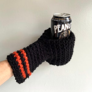 This beer mitten is black with orange gold stripes around the wrist. It is part crochet can holder part mitten. A hand is in it and a can of beer inside the holder. The bottom is covered so the can stays in place. It can be used on either hand.