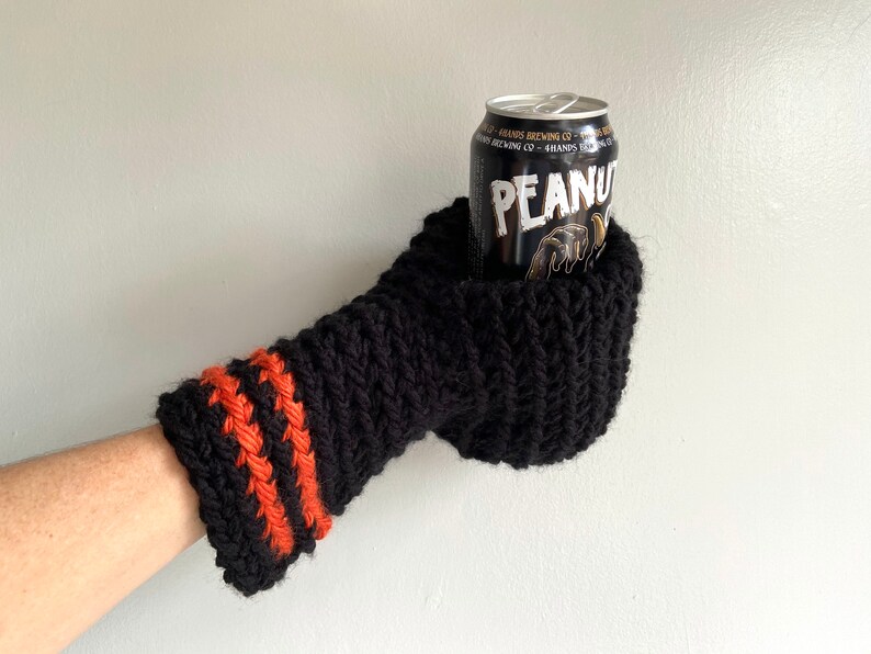 This beer mitten is black with orange gold stripes around the wrist. It is part crochet can holder part mitten. A hand is in it and a can of beer inside the holder. The bottom is covered so the can stays in place. It can be used on either hand.