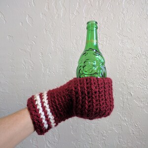 This beer mitten is red with white stripes around the wrist. It is part crochet can holder part mitten. A hand is in it and a can of beer inside the holder. The bottom is covered so the can stays in place. It can be used on either hand.