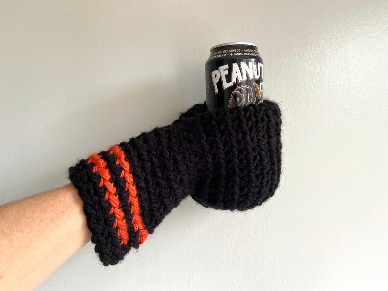 This beer mitten is black with orange gold stripes around the wrist. It is part crochet can holder part mitten. A hand is in it and a can of beer inside the holder. The bottom is covered so the can stays in place. It can be used on either hand.