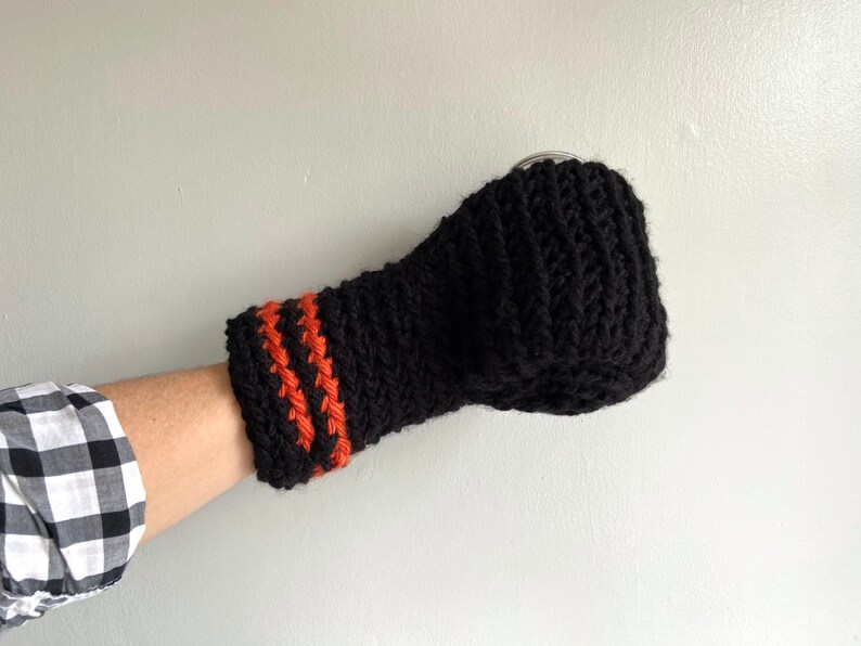 This beer mitten is black with orange gold stripes around the wrist. It is part crochet can holder part mitten. A hand is in it and a can of beer inside the holder. The bottom is covered so the can stays in place. It can be used on either hand.