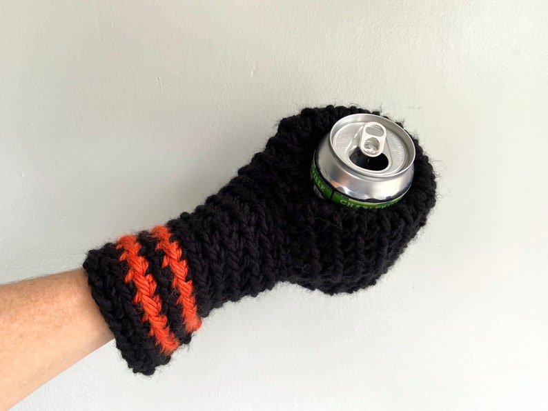 This beer mitten is black with orange gold stripes around the wrist. It is part crochet can holder part mitten. A hand is in it and a can of beer inside the holder. The bottom is covered so the can stays in place. It can be used on either hand.