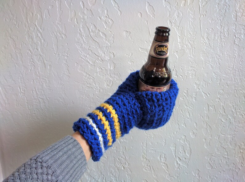This beer mitten is blue with white and yellow stripes around the wrist. It is part crochet can holder part mitten. A hand is in it and a can of beer inside the holder. The bottom is covered so the can stays in place. It can be used on either hand.