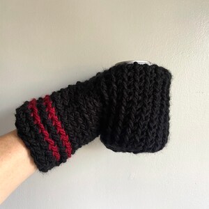 This beer mitten is black with red stripes around the wrist. It is part crochet can holder part mitten. A hand is in it and a can of beer inside the holder. The bottom is covered so the can stays in place. It can be used on either hand.