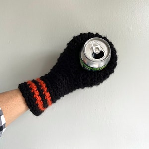 This beer mitten is black with orange gold stripes around the wrist. It is part crochet can holder part mitten. A hand is in it and a can of beer inside the holder. The bottom is covered so the can stays in place. It can be used on either hand.