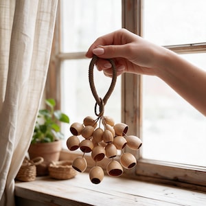 Puede incluir: Un adorno colgante decorativo que presenta un grupo de pequeñas piezas de madera de color claro en forma de campana, lisas, suspendidas de un anillo de metal central unido a un lazo de cuerda trenzada color canela.