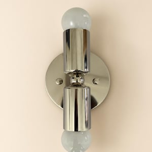 Pode incluir: Um candeeiro de parede com uma placa circular de níquel polido e dois abajures metálicos cilíndricos verticais. Cada abajur é rematado com uma pequena lâmpada redonda de vidro branco fosco no topo e na base, criando um design simétrico.