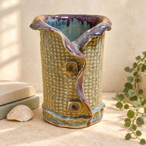 May include: A cylindrical ceramic vase with a textured, grid-like surface in shades of olive green and tan. The rim and a vertical, folded-over accent feature a glossy, deep purple and blue glaze, with two small square indentations on the side.