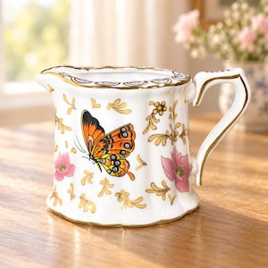 Peut inclure: Un pot à lait en porcelaine blanche orné d'un motif de papillon orange et noir, agrémenté d'illustrations florales roses et de motifs en filigrane dorés. La pièce présente un bord festonné et une anse dorée, avec une finition brillante.