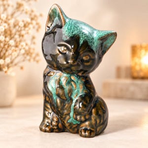 May include: A ceramic cat figurine featuring a glossy dark brown finish with vibrant turquoise green glaze accents dripping from the ears and chest. The cat is depicted in a seated position with its head tilted slightly to the side.