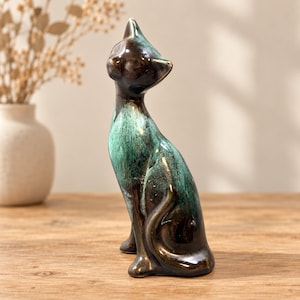 May include: A ceramic cat figurine in a seated pose with its head tilted upward. The sculpture features a glossy finish with a color gradient transitioning from deep dark brown at the head and base to a vibrant teal green across the chest and back.
