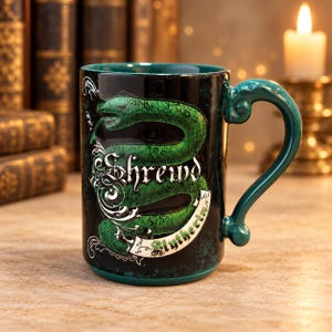 Puede incluir: Una taza de cerámica negra con un gráfico de una serpiente verde enroscada y el texto Shrewd y Slytherin. La taza tiene un interior verde oscuro y un asa curva. El diseño incluye detalles de filigrana plateada y una textura agrietada.