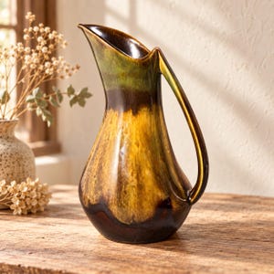 May include: A ceramic pitcher featuring a glossy glaze with a gradient of deep brown at the base, transitioning into a vibrant mustard yellow in the centre, and finishing with a dark olive green at the rim, complete with a curved handle.