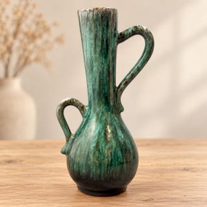 May include: A ceramic vase featuring a bulbous base and a tall, slender neck with two handles of varying sizes. The surface is finished with a glossy glaze in shades of deep forest green, teal, and dark brown, creating a vertical drip pattern.