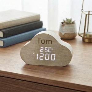 May include: A cloud-shaped wooden digital clock featuring the name Tom engraved on the front. The display shows the time as 12:00 and the temperature as 25C in bright white digital numbers against the light wood grain surface.