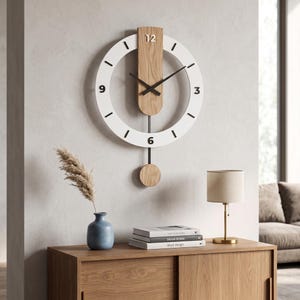 Könnte beinhalten: Eine runde Wanduhr mit weißem Rahmen, schwarzen Stundenmarkierungen und den Ziffern 3, 6, 9 und 12. Ein vertikales helles Holzpaneel in der Mitte trägt schwarze Zeiger und eine dünne Pendelstange mit einem runden Gewicht aus Holz.