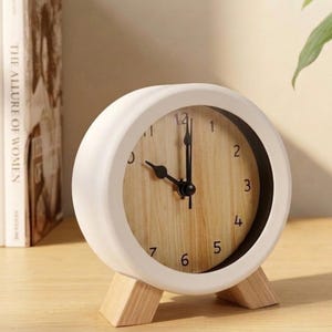 May include: A round analog table clock with a white frame and two light wood-toned feet. The clock face features a light wood grain pattern with black numerals from 1 to 12 and black hour, minute, and second hands pointing to approximately 9:09.