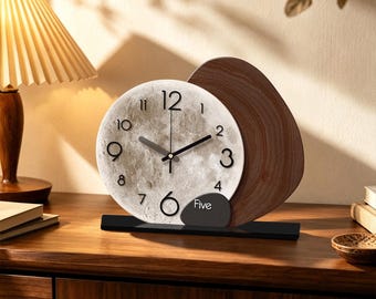 May include: A table clock featuring a circular face with a textured moon surface pattern, black numerals, and black hands. It is partially overlapped by a dark brown wood-grain panel and a small black accent piece with the word Five printed on it.