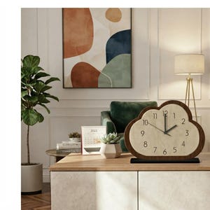 May include: A cloud-shaped analog table clock with a dark wood frame and a light-colored face featuring black numerals from one to twelve. The clock has two black hands and sits on a rectangular black base, positioned on a light-colored surface.