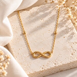 May include: A gold-toned necklace featuring a central infinity symbol pendant. The chain is composed of delicate links interspersed with small, smooth spherical beads. The metal has a polished, reflective finish, creating a bright appearance.