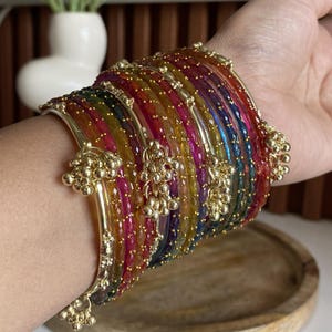 May include: A stack of multi-coloured translucent bangles featuring a faceted texture. Each bangle is accented with small gold-toned metal beads and dangling gold-toned charms. The set includes various vibrant hues such as red, yellow, blue, and green.