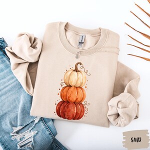 May include: A sand-colored crewneck sweatshirt featuring a graphic print of three stacked pumpkins in shades of cream, orange, and dark red. The sweatshirt has long sleeves and a ribbed collar, paired with distressed blue denim jeans.