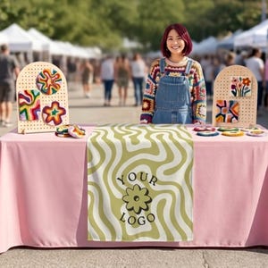 May include: A pink tablecloth featuring a central runner with a wavy olive green pattern and the text "YOUR LOGO" above a flower icon. Two arched pegboards display colorful tufted fiber art pieces in various shapes, including flowers and abstract swirls.