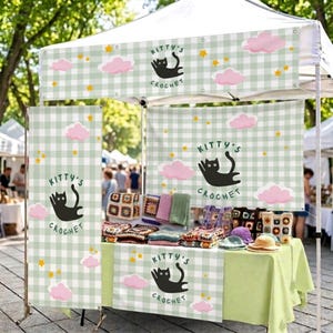 May include: A vendor booth featuring a light green and white gingham canopy and banners with the text Kitty's Crochet, black cat illustrations, pink clouds, and yellow stars. A table displays various colorful crocheted items and hats.
