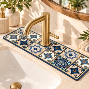 Puede incluir: Una alfombrilla absorbente rectangular para fregadero con un patrón repetitivo de azulejos florales en azul, blanco y dorado. La alfombrilla se coloca alrededor de la base de un grifo de oro cepillado sobre una encimera clara junto a un lavabo blanco.