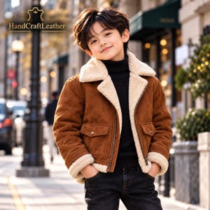 Könnte beinhalten: Eine braune Wildlederjacke mit cremefarbenem Kunstfellfutter, einem breiten Kragen und zwei Vordertaschen mit Klappen und Metallknöpfen. Die Jacke hat einen durchgehenden Reißverschluss und wird über einem schwarzen Rollkragenpullover getragen. Text: HandCraftLeather.
