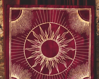 Puede incluir: Un cuadrado de tela roja con un diseño de sol dorado. El sol tiene un círculo grande en el centro y rayos que se extienden hacia afuera. La tela tiene una textura sutil.